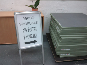 Mittig steht ein Aufsteller mit der Aufschrift: Aikido Shofukan. Im Hintergrund ist eine Komode, mit einer kleinen Pflanze drauf, zu sehen und rechter Hand ein Stapel Aikidomatten.
